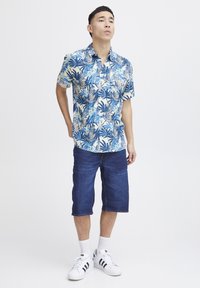 Camicia a maniche corte, blu con motivo floreale in verde e bianco, abbinata a pantaloni corti in denim blu scuro e sneakers bianche con strisce nere.