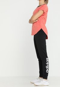 Roze T-shirt met korte mouwen en een grijze halslijn, gecombineerd met zwarte sweatpants met witte "adidas" branding. Witte sportieve schoenen zichtbaar.