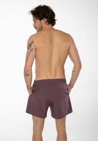 Pantaloni da bagno viola con cintura regolabile, dettaglio tasca sul retro e una texture liscia. Il modello mostra la vista posteriore.