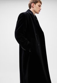Μαύρο βελούδινο παλτό διπλής όψης με όχι κολάρο, πλαϊνές τσέπες και κουμπωτές λεπτομέρειες στους μανίκια. Λείος υφασμάτινος υφής και ραμμένη εφαρμογή.
