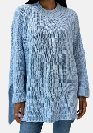 Kvinde iført løs lyseblå strikket sweater med faldende skuldre, opsmøgede ærmer og sideslidser over blå jeans, kort ved halsen og midt på låret.