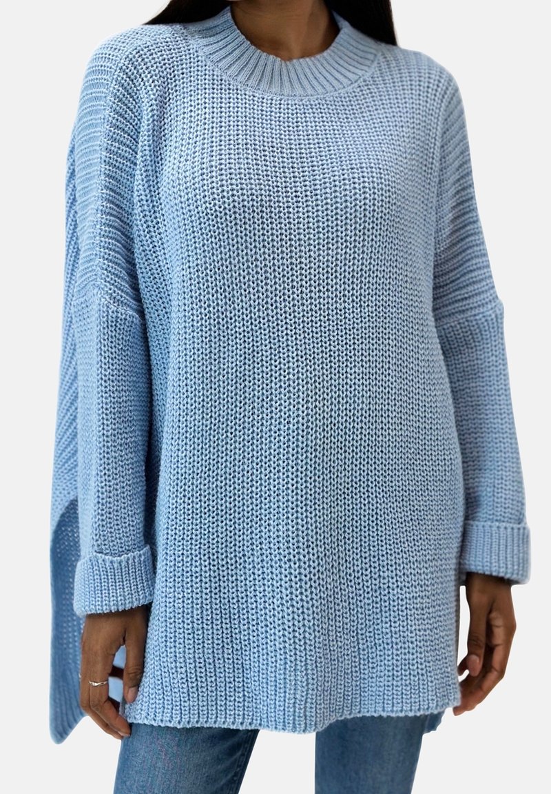 Femme portant un pull tricoté ample bleu clair avec épaules tombantes, poignets roulés et fentes latérales sur un jean bleu, raccourci au niveau du cou et à mi-cuisse.