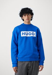 Blauwe sweatshirt met een witte "HUGO" logo over de borst, ronde hals en lange mouwen. Voorzien van geribbelde boorden en onderrand. Model draagt grijze broek.