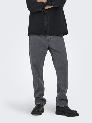 Person wearing loose gray denim jeans, black leather loafers, and a black button-up shirt with arms crossed.