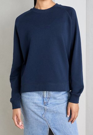 Kvinde iført en mørkeblå langærmet crewneck-sweatshirt og et lyseblåt denimsnørk med en forreste slidse.