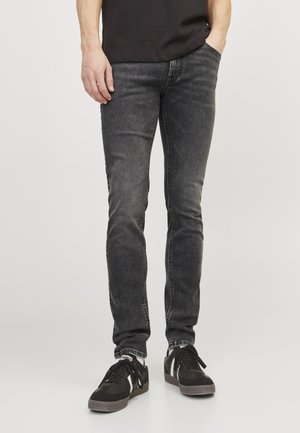 Mann trägt schlank geschnittene ausgewaschene schwarze Jeans mit schwarzen Sneakers und einem schwarzen T-Shirt, steht mit einer Hand in der Tasche.