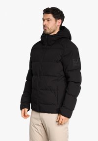 Doudoune noire avec capuche, dotée d'une fermeture éclair sur le devant et de poignets élastiques. Design matelassé notable et un patch logo sur le bras gauche.