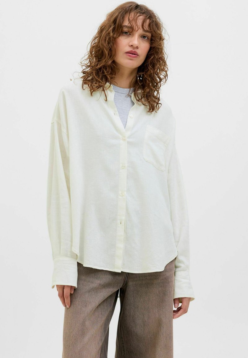 Camicia button-up gialla chiara con taschino, maniche lunghe e vestibilità rilassata, abbinata a pantaloni a gamba larga marroni. Texture liscia.