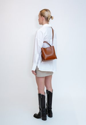 Femme aux cheveux blonds en chignon portant une chemise blanche, une jupe beige, des bottes noires montantes et un sac à dos en cuir marron sur une épaule.