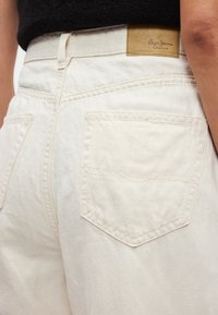 Bílé džínové kalhoty s dvěma zadními kapsami, viditelným prošíváním a značkovou hnědou koženou nášivkou na pase. Evidentní matná textura.