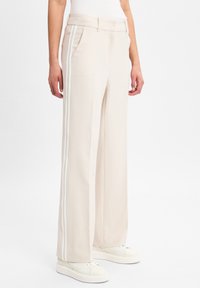 Wijde broek gemaakt van een lichtbeige stof, met witte zijstrepen, voorzakken en een gladde textuur. Gestyled met witte sneakers.