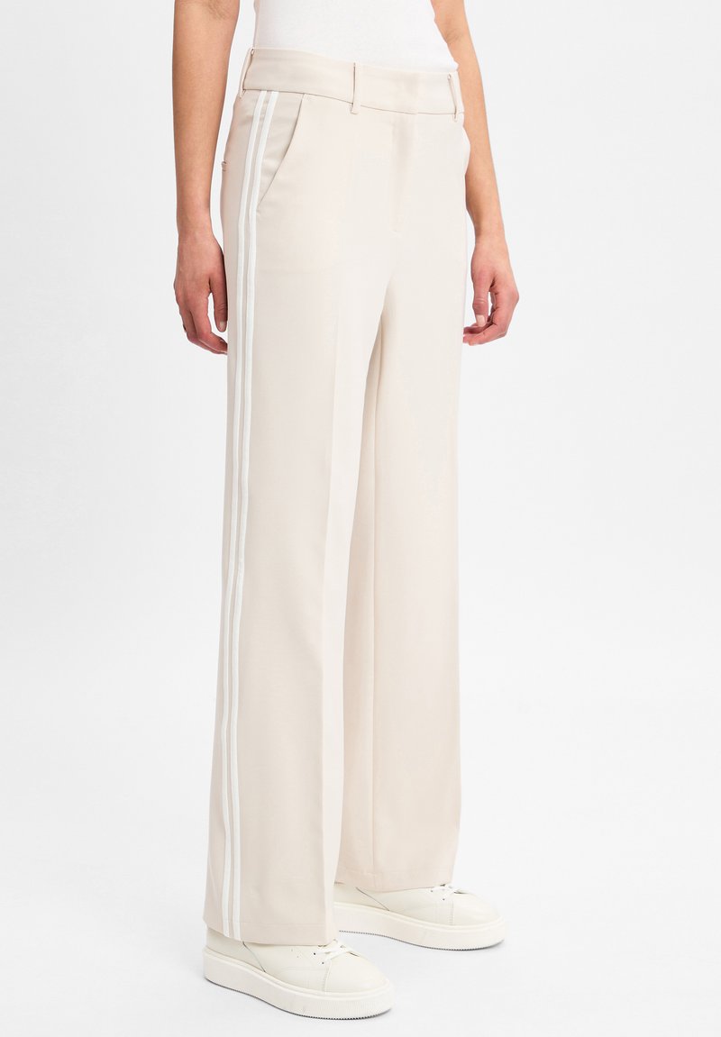 Wijde broek gemaakt van een lichtbeige stof, met witte zijstrepen, voorzakken en een gladde textuur. Gestyled met witte sneakers.