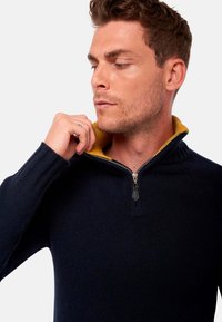 Pull en tricot marine avec un col jaune, fermeture zip, poignets côtelés et finition texturée. Dispose d'un design de col montant.