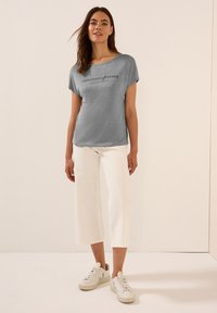 Grijs T-shirt met korte mouwen van reflecterend materiaal en de tekst "CRESCENDO COLORS". Gecombineerd met witte wijde broek en lichte sneakers.
