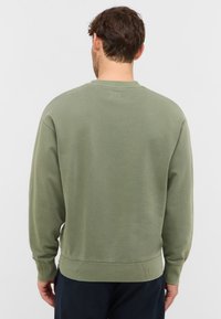 Homme avec des cheveux bruns courts portant un sweat-shirt à manches longues vert olive et un pantalon foncé, se tenant de dos devant un fond blanc uni.