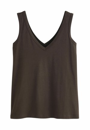 Femme portant un blazer bordeaux et un pantalon à jambes larges avec un haut noir à col en V, debout les mains dans les poches devant un fond beige et blanc.