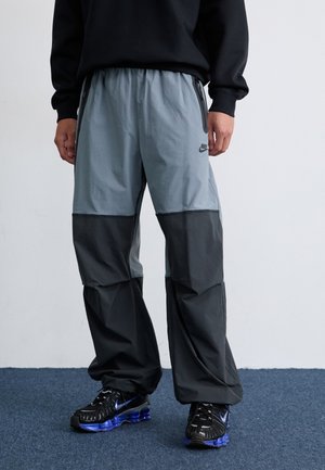 Een persoon staat in een zwart sweatshirt en grijze Nike sportbroek met zwarte accenten en zwarte sneakers, tegen een lichte achtergrond.