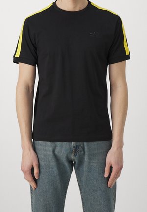 Mann trägt ein schwarzes Kurzarmshirt mit gelben Streifen auf den Schultern und blaue Jeans, steht vor einem schlichten hellen Hintergrund.