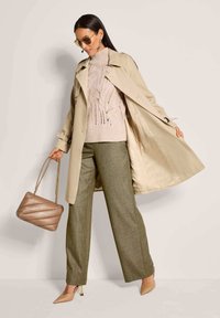 Beige trenchcoat over een lichtroze gebreide trui met bladpatronen, groene wijde broek en een gewatteerde beige handtas.