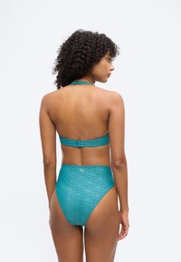 Γυναίκα με σγουρά μαλλιά που φοράει ένα πράσινο μπικίνι δύο τεμαχίων με μαγιό με ψηλή μέση και λογότυπα Puma, εμφανιζόμενη από πίσω σε απλό φόντο.