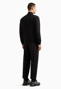 Tuta sportivo nero con giacca zip-up e pantaloni. Tessuto morbido, forma aderente e logo EA7 visibile sul retro della giacca.