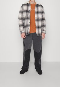 Giacca a quadri bianca e nera con colletto in shearling, felpa arancione e pantaloni larghi grigi con toppe curve. Scarpe nere completano l'outfit.
