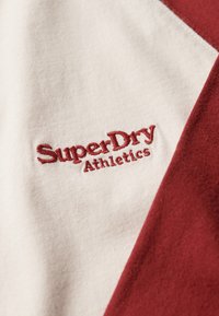 Rood en crèmekleurige katoenen stof met een hoekig ontwerp. Heeft een geborduurd "SuperDry Athletics" logo in bordeaux. Zachte textuur is evident.