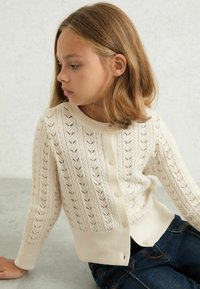 Cardigan bianco lavorato a maglia con un motivo a foglia, collo rotondo e bottoni sul davanti. Ha polsini e orlo a coste, abbinato a jeans scuri.
