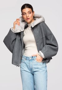 Femme ajustant une veste en laine grise doublée en duvet, portant un col roulé beige et un jean bleu clair, posant devant un fond uni clair.