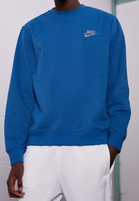 Felpa blu a girocollo realizzata in un tessuto misto cotone. Presenta un logo Nike argentato sul lato sinistro del petto e polsini e orlo a costine.