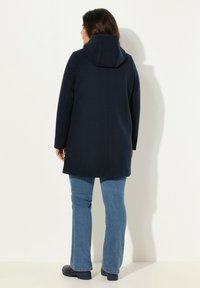 Cappotto blu navy con cappuccio, realizzato in tessuto morbido. Presenta un design pulito, una singola cucitura lungo la schiena ed è lungo fino al ginocchio.