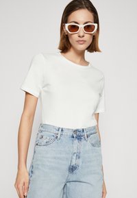 Weißes T-Shirt mit kurzen Ärmeln und Rundhalsausschnitt, kombiniert mit hellblauen, hochgeschnittenen Jeans. Das Model trägt eine braun getönte Sonnenbrille.