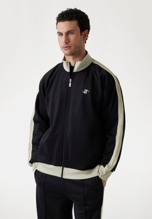 Giacca nera con zip e colletto alto, con strisce laterali color crema e polsini a coste. Logo visibile sul petto. Tessuto liscio.