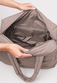 Ręce trzymają otwartą dużą torbę w odcieniu taupe z wyściełanymi uchwytami, ukazując pustą wnętrze z zamykaną kieszenią oraz metką z materiału zrównoważonego rozwoju.