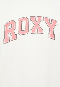 Fehér póló nagy, feltűnő, rózsaszín "ROXY" grafikával. A szöveg barna kontúrral rendelkezik, ami éles kontrasztot teremt az anyaggal.