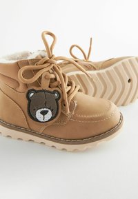 Hnedé boots z faux kože s kožušinovým podšívkou, nášivkou hnedého medveďa a textúrovanou gumovou podrážkou. Obsahujú šnúrky a kontrastné prešívanie.