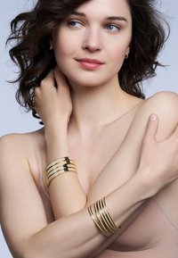 Des bracelets en métal doré présentent un design empilé avec des surfaces lisses et brillantes ainsi que des rayures fines et arrondies. Le modèle porte un haut neutre.