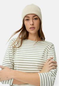 Cremefarvet strikket hue med ribbet tekstur; person iført stribet bluse i beige og off-white, med armene over kors, neutral baggrund.