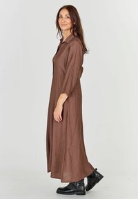 Brown, long-sleeve maxi dress made of textured fabric, featuring a collar and side slits, paired with black ankle boots for contrast.