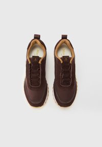 Une paire de baskets marron foncé avec un intérieur beige et des tirettes, présentant un dessus lisse et des surpiqûres, sur fond blanc.