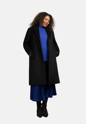 Zwarte wolmix jas met een geknipte kraag en twee voorzakken, gedragen over een levendig blauwe trui en een geplooide blauwe rok. Zwarte puntige laarzen.
