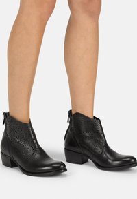 Botines de cuero negro con diseño perforado, parte superior curvada y tacón bajo apilado; cremalleras en la parte trasera para facilitar su uso.