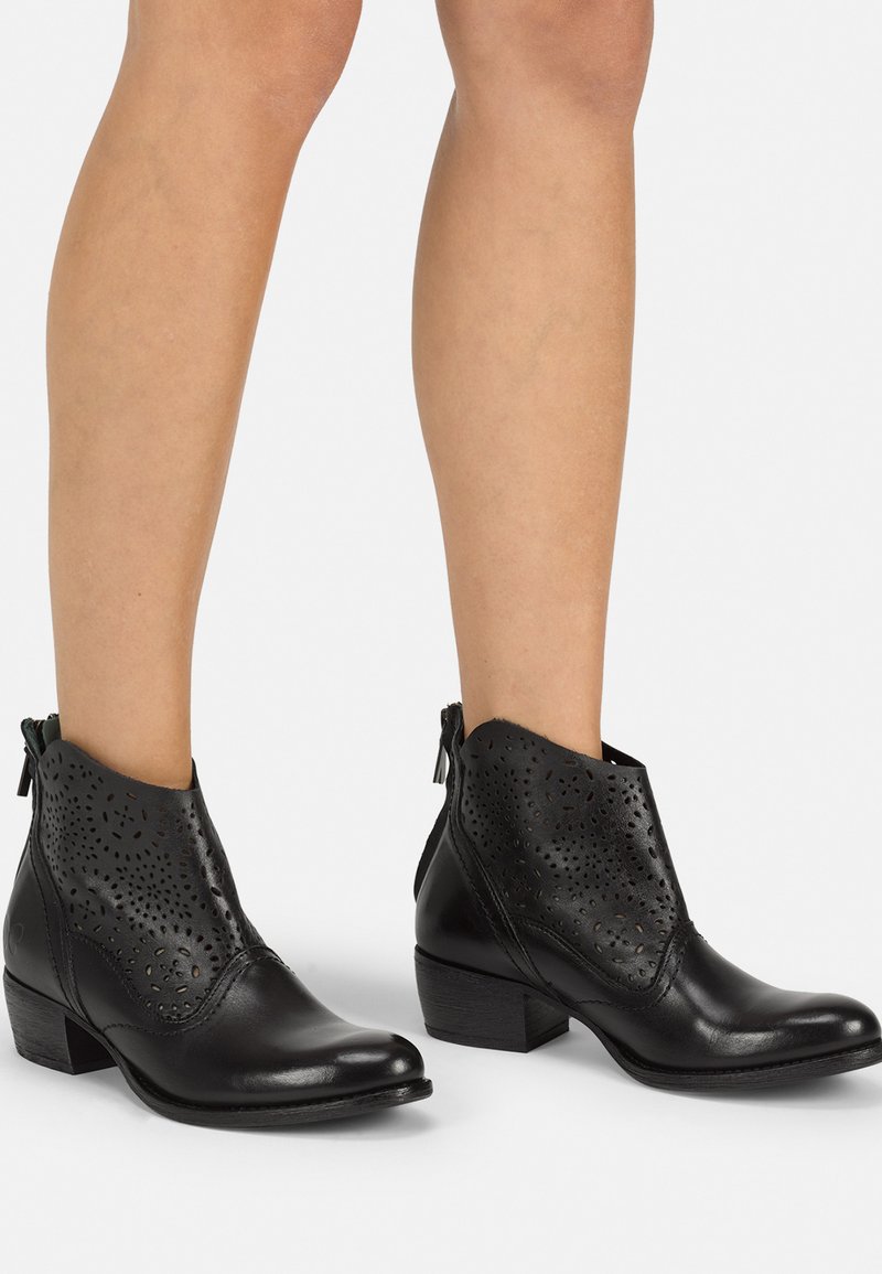 Botines de cuero negro con diseño perforado, parte superior curvada y tacón bajo apilado; cremalleras en la parte trasera para facilitar su uso.