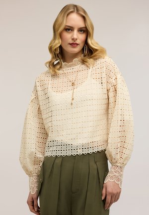 Vaaleahiuksinen nainen, joka on pukeutunut kermanväriseen pitsipaitaan, jossa on aallotetut reunat ja pitkät pussihihat, yhdistettynä oliivinvihreisiin korkeavyötäröisiin housuihin.