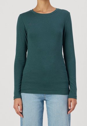 Frau trägt ein tailliertes, langärmeliges Shirt in Türkis und hellblaue Jeans, gezeigt von Hals bis Hüfte vor einem schlichten Hintergrund.