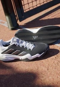 Sort-hvide Adidas tennissko på en grusbane tæt på tennisnetstolpen.