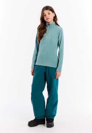 Jong persoon staat in een teal geribbelde long-sleeve pullover, losse teal broek en zwarte platformlaarzen tegen een witte achtergrond.