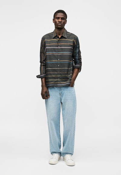 Άνδρας που στέκεται με χέρια στις τσέπες, φοράει σκούρο ριγέ πουκάμισο με κουμπιά, ανοιχτό μπλε τζιν και λευκά αθλητικά παπούτσια σε απλό φόντο.