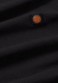 Čierna látka s viditeľnou textúrou, obsahujúca malý štvorcový záplatu s vyšívaným oranžovým motívom pripomínajúcim slnko. Hladký povrch bez výrazných vzorov.