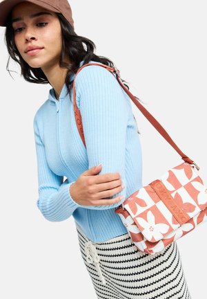 Mujer con suéter de canalé azul claro y falda a rayas, que lleva una gorra marrón y un bolso de hombro blanco con estampado floral en tonos terracota.